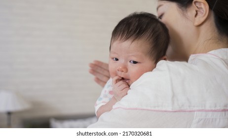 selective focus cute newborn infant is looking at camera innocently as her mommy is helping her burp at blurred background bright nursery room.