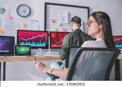Selective focus of data analyst holding pen near colleague working with computers at table