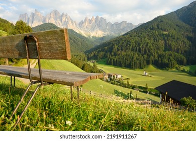 (Selective focus) Defocused park bench in the foreground with the Church of St. John (San Giovanni in Ranui) that stands out in the green meadows, in the heart of the beautiful Dolomitic landscape.	