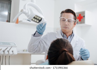 Selective Focus Of Dentist In Face Shield And Latex Gloves Touching Medical Lamp Near Woman