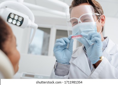 Selective Focus Of Dentist In Medical Mask, Face Shield And Latex Gloves Holding Dental Prosthesis Near African American Woman