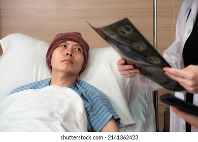 Selective Focus Of A Depressed Asian Male Cancer Patient In A Head Cover Laying On Bed Looking At A Blurred And Cropped Doctor Who Is Diagnosing His Brain MRI Scan Film To The Nurse Beside His Bed.