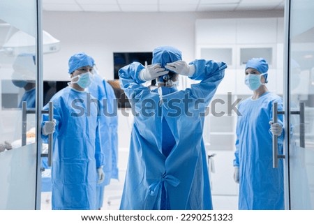 Selective focus of a doctor's back in surgical uniform and gloves, tying a surgical cap while walking into an operating room with two medical assistants in surgical clothes standing holding door open.