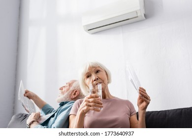 Selective focus of elderly woman holding glass of water and fan near husband suffering from heat at home