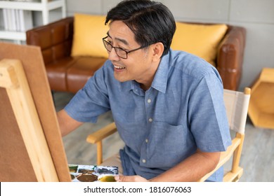 Selective Focus At Eye And Glasses. Indoor Shot Of Senior Asian Man Sit And Painting Picture Inside Of The House. Happy Retirement Lifestyle Concept.