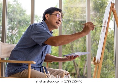 Selective Focus At Face. Indoor Shot Of Senior Asian Man Sit And Painting Picture Inside Of The House. Happy Retirement Lifestyle Concept. 