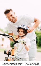 Selective Focus Of Father And Son Looking At Camera While Dad Putting Helmet On Boy 