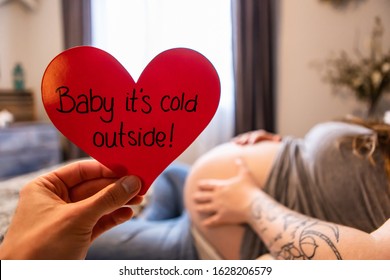 A Selective Focus First Person Perspective Of A Hand Holding A Red Heart With Message Baby Its Cold Outside, As Heavily Pregnant Woman Rests In Bed