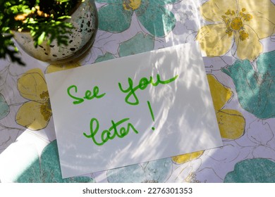 Selective focus flat lay view of table with small mercury glass vase and the message See You Later on card set on pretty flowered paper in dappled light