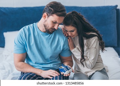 Selective Focus Of Grieving Parents Sitting On Bed And Holding Baby Shoes At Home