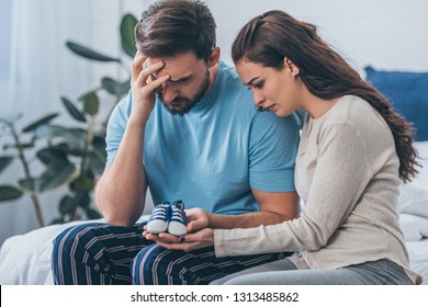 Selective Focus Of Grieving Parents Sitting On Bed And Holding Baby Shoes At Home