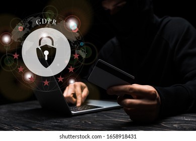 selective focus of hacker in mask using laptop while holding credit card near padlock with gdpr lettering on black