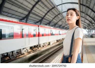 Selective focus half-body image of a beautiful Asian female traveler standing waiting for an arrival of skytrain on a platform with a blurred train in opposite side as a background at skytrain station
