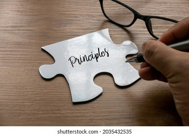 Selective focus of a hand writing word ' Principles ' on a a piece of white jigsaw puzzle with a glasses on wooden background. - Shutterstock ID 2035432535