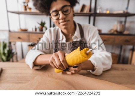 selective focus of handmade candle in hands of positive african american woman in eyeglasses and apron on blurred background
