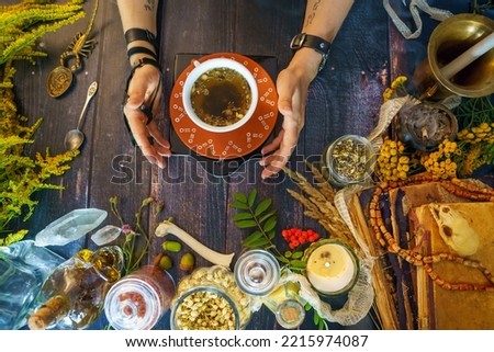 Selective focus hands close to a cup of warm tea. Brewed herbs in a ceramic pot on the wooden table . Magical Tea Recipes for witches. Teacup with herbs for ritual cleansing. A special blend of plants