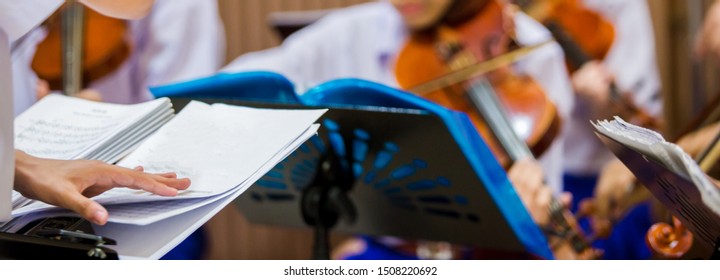 Selective Focus To Hands Of Conductor With Music Notation. Hands Of  Conductor With Blurry Asian Boy Students Playing Violin With Music Notation In The Group. Symphony Orchestra On Stage.