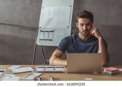 Selective Focus Of Handsome Ux Designer Looking At Camera Beside Laptop And Sketches On Table