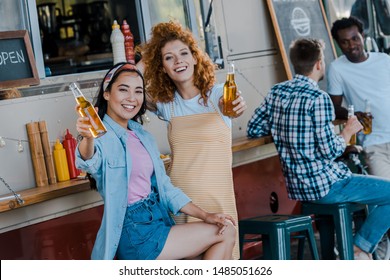 selective focus of happy asian girl holding beer near redhead woman and food truck