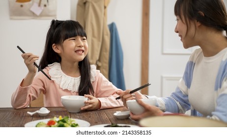 Selective Focus Of Happy Asian Girl Having Yummy Vegetable With Satisfied Face. She Tells Her Mom She Likes The Food As The Woman Pointing At Another Dish On Dinner Table