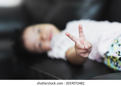 Selective focus. Happy blur child pointing up two fingers. Asian girl pointing number two fingers sign, v for victory hand gesture. Kid lying down on sofa. 
