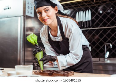 selective focus of happy chocolatier in black latex gloves holding pastry bag near chocolate molds