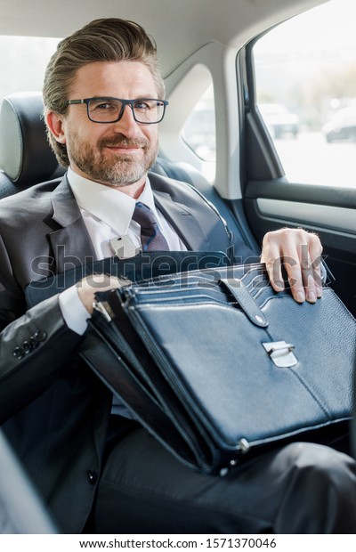 selective focus of happy diplomat in glasses holding\
briefcase in car