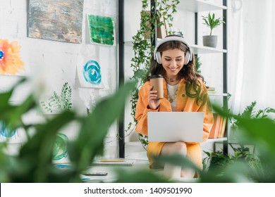 Selective Focus Of Happy Young Woman With Paper Cup Listening Music In Headphones And Using Laptop