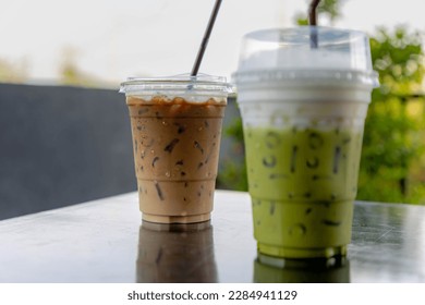 Selective focus of iced matcha with white foam milk on top and iced espresso, Takeaway homemade cold drink in plastic glass and straw on the table with blurred green garden background.