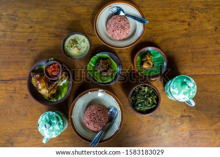 Selective focus image flat lay set of Sego Abang Gunung Kidul or Javanese food set contains of red rice, beef stew, chicken liver, tempeh with green chilli like soup, sauted papaya leaf.
