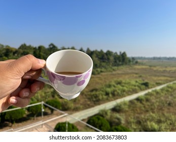 Selective Focus Image Of A Male Hand Holding A Half Empty Cup Of Tea Against Green Fields And Blue Sky Background On A South Asian Village.