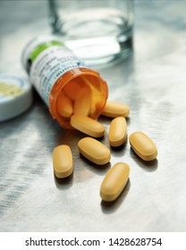 Selective Focus Image Of Yellow Capsules Spilling From Prescription Medication Bottle With Glass Of Water On Steel Counter