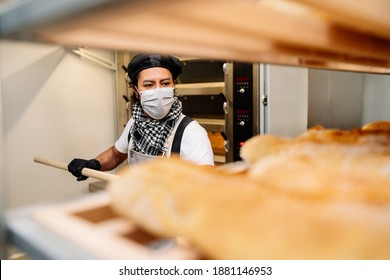 Selective Focus Of Latino Baker Shelving Freshly Baked Bread With A Protective Face Mask For The 2020 Covid 19 Coronavirus Pandemic