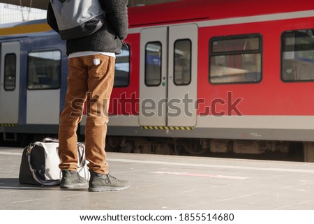 Selective focus, low angle view of male traveler with his backpack and shoulder bag who wear winter jacket and Corduroy trouser wait for arrival train on the platform railway station.