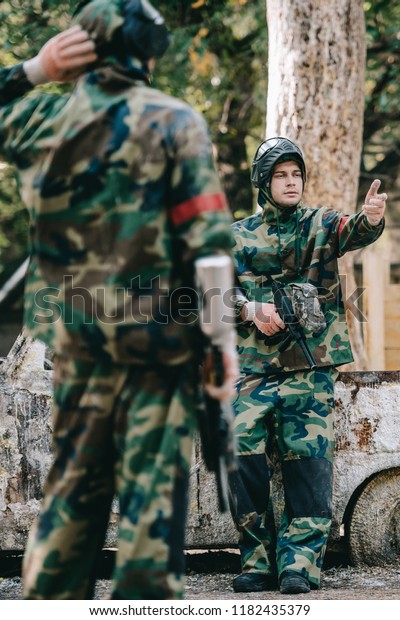 selective focus of male paintballer in camouflage
uniform holding marker gun and pointing by hand to his team near
broken car outdoors 