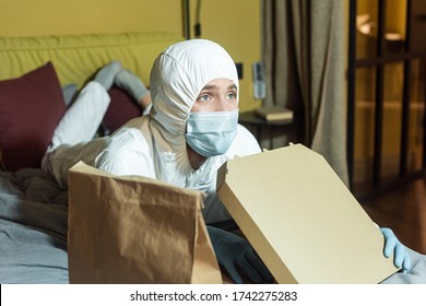 Selective Focus Of Man In Medical Mask And Hazmat Suit Watching Tv On Bed Near Package And Pizza Box