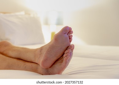 Selective Focus Of A Men Feet Lied Down On Bed, Relaxing And Watching TV On White Bed Sheet, A Man Showing His Bare Foot On Blanket With Lamp Light At Night In Bedroom.