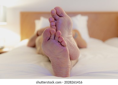 Selective Focus Of A Men Feet Lied Down On Bed, Relaxing And Watching TV On White Bed Sheet, A Man Showing His Bare Foot On Blanket With Lamp Light At Night In Bedroom.