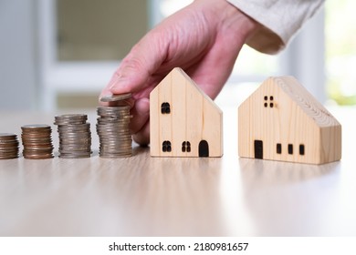 Selective Focus Men Hand Putting Coin On Stack Of Money Coin For Saving For Home, Property With Small Wood House On The Background. Wealth And Saving Plan For House Mortgage, Personal Investment.