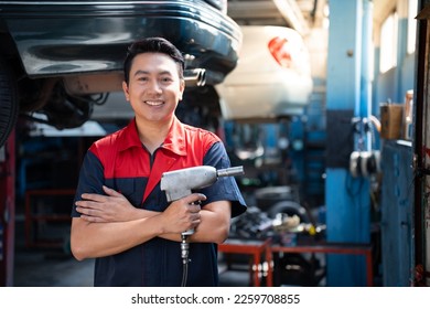 Selective focus of mid-adult Asian male mechanic in red blue uniform, standing hold an impact wrench with arms folded and smiling at camera with blurred garage in background. Copy space on the right. - Powered by Shutterstock