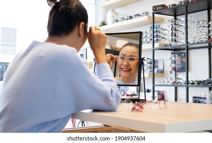 Selective focus at the mirror.  Senior elder Asian women smile and look at the mirror while choosing beautiful glasses frame trial inside of optical shop, store.  Eye sight for elder, healthcare.