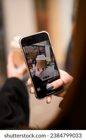 Selective focus of mobile screen with photo of paper cup takeaway coffee decorated with rose flower in female hand. Female hand taking picture of paper coffee cup