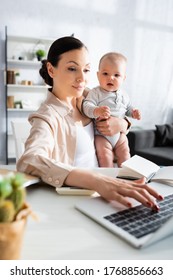 selective focus of mother typing on laptop keyboard and holding in arms baby boy
