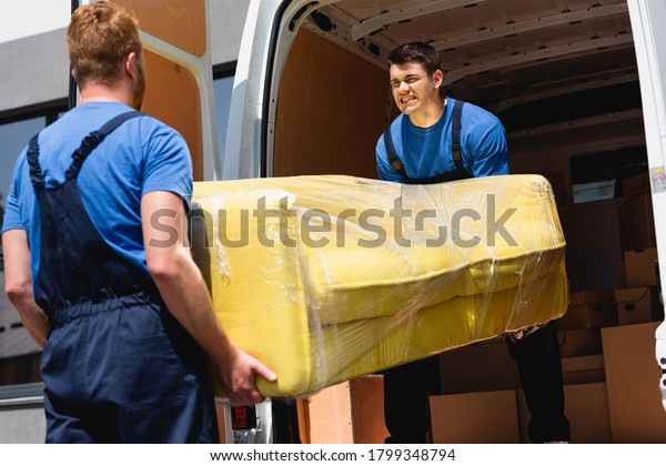 Selective focus of movers unloading sofa in\
stretch wrap in truck\
outdoors