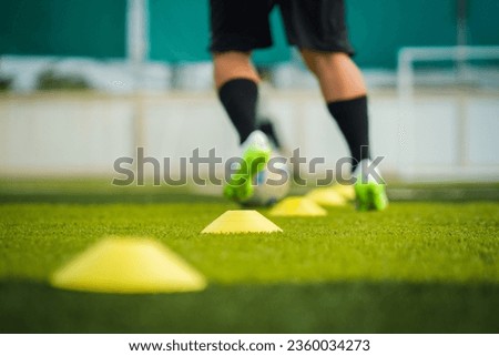 Selective focus at obstacle cones which are placed on turf pitch ground with blurred of a football player is dribbling the ball for training. Sport training in action and backgorund photo.
