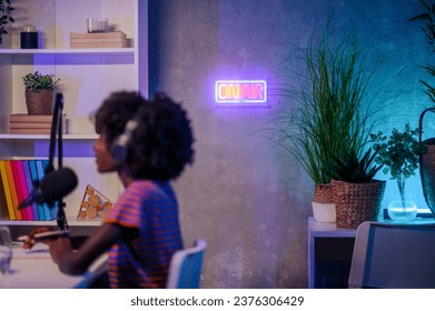 Selective focus on the "on air" neon sign in a small home broadcasting and recording studio with a podcaster in a blurry foreground making a podcast. Radio object in a podcasting studio. - Powered by Shutterstock