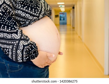 Selective Focus On Anonymous Woman Pregnant Belly In Birth Clinic, Out Of Focus Hospital Corridor On The Background, Regular Medical Checkups Concept.