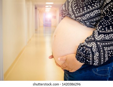 Selective Focus On Anonymous Woman Pregnant Belly In Birth Clinic, Out Of Focus Hospital Corridor On The Background, Regular Medical Checkups Concept.