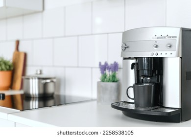 selective focus on automatic espresso machine with dark mug on white countertop at stylish kitchen with household appliances and kitchenware on blurred background