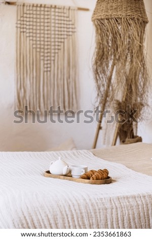 Selective focus on bamboo tray with teapot, cup and croissants on soft cotton bedding. Tasty breakfast in bed in lifght room with decor elements.